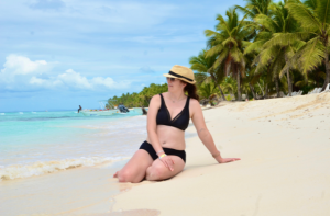 Louise sitting on the sand on a Caribbean beach in Punta Cana, looking out at the turquoise sea with palm trees and boats in the background.